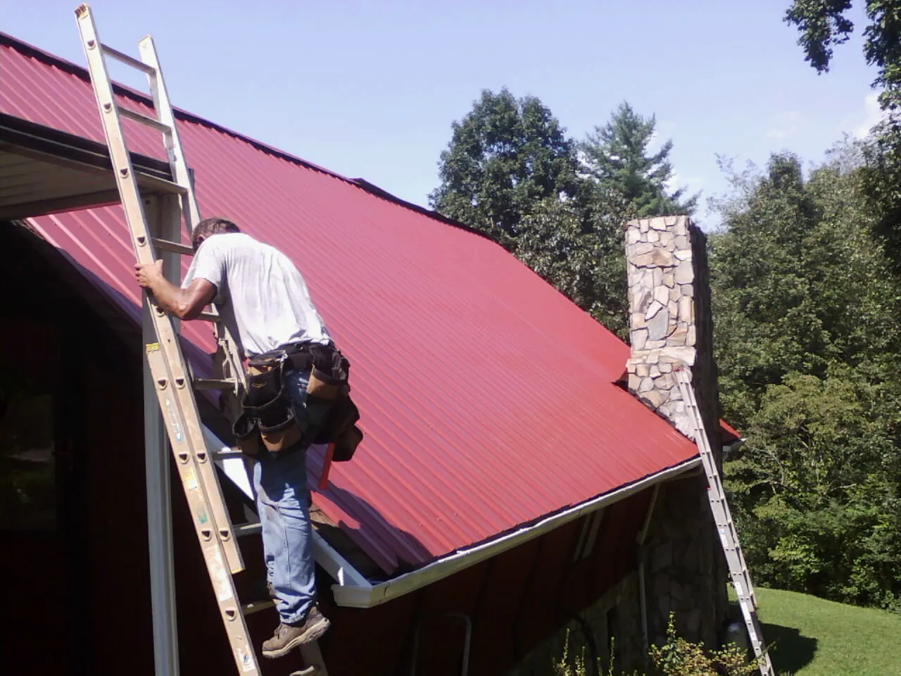 Roofer on a ladder beside a red metal roof and stone chimney — Soffit & Fascia Repair by Roofing Brothers in Greenville